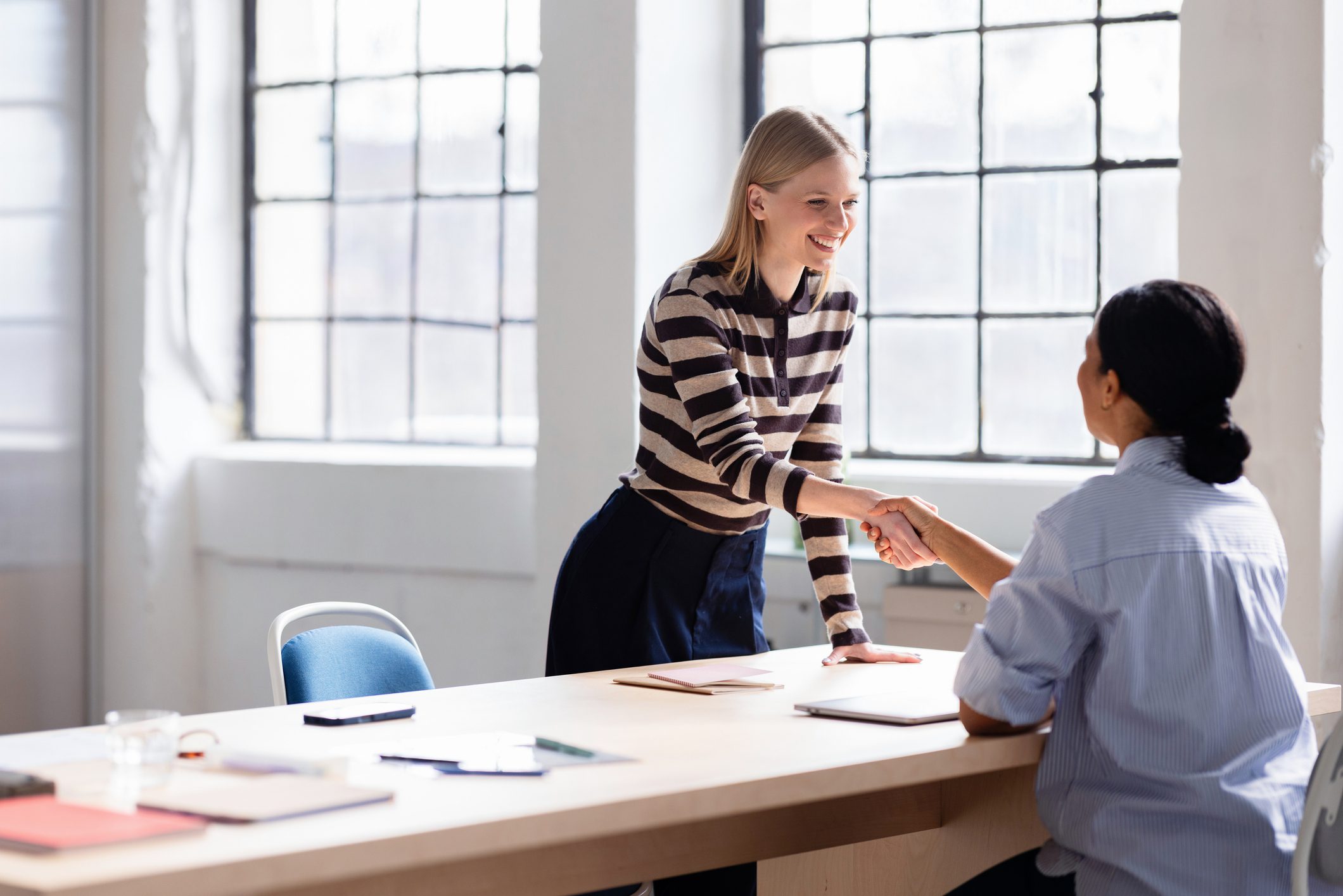 Two professional women shake hands across a bright office table, conveying agreement and positive collaboration.